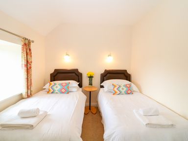 A room with two single beds each with white bedding and colorful pillows with a round table in-between holding a vase of yellow flowers at Cornflower Cottage in St Columb Major