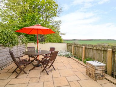 An outdoor patio with a wooden table and chairs under a red umbrella and a view of fields at Cornflower Cottage in St Columb Major