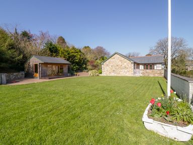 A grassy yard with a small wooden shed stone building and a flower bed with red flowers at Cornflower Cottage in St Columb Major