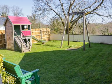 A garden with a wooden playhouse with slide a swing set and a green bench at Forget-me-not in St Columb Major