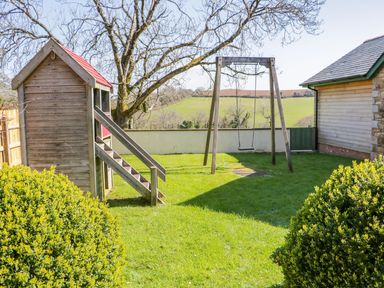 A backyard with a wooden playhouse and a swing set with a green lawn at Forget-me-not in St Columb Major