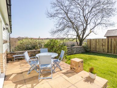An outdoor patio with a round table and four chairs next to a lawn with a tree and a wooden fence at Forget-me-not in St Columb Major