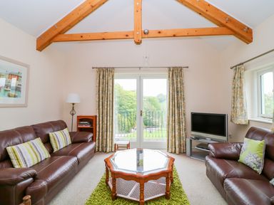 A living room with leather sofas and a coffee table facing glass doors with curtains at Forget-me-not in St Columb Major
