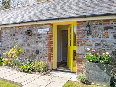 An entrance with a yellow door in a stone building with flowers in a planter and garden bed at FernGully in St Columb Major