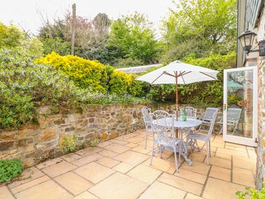 A stone patio with a white metal table and chairs under a white umbrella near a stone wall and greenery at FernGully in St Columb Major
