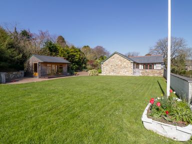 A garden with a large lawn a wooden shed a stone building and a flower bed with red tulips at FernGully in St Columb Major