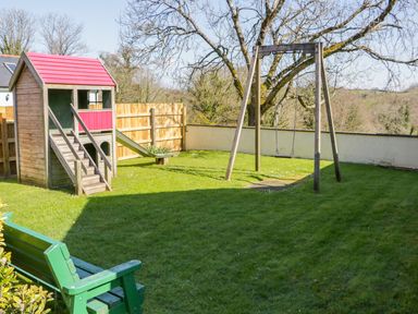 A garden with a wooden playhouse with slide a swing set and a green bench at Kingcup Cottage in St Columb Major