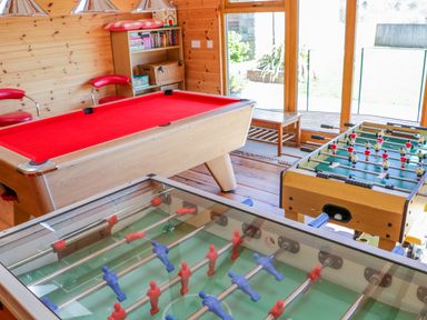 A game room with a red pool table two foosball tables wooden shelves with books and chairs at Kingcup Cottage in St Columb Major