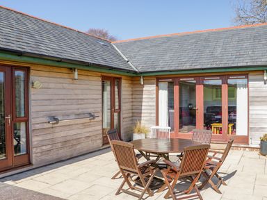 An outdoor patio area with a round wooden table and six chairs outside a building with wooden siding and glass doors at Kingcup Cottage in St Columb Major