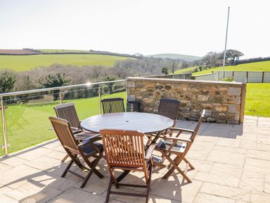 An outdoor patio with a round wooden table and six wooden chairs overlooking green fields at Kingcup Cottage in St Columb Major