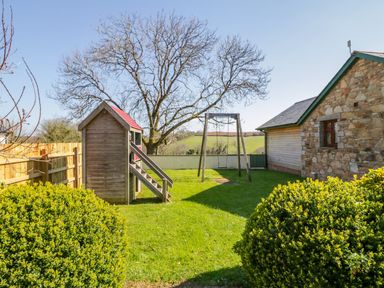 A garden with a wooden playhouse and swing next to a stone and wood building at Kingcup Cottage in St Columb Major