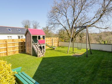 A backyard with a wooden playhouse with a red roof a swing set and green grass at Kingcup Cottage in St Columb Major