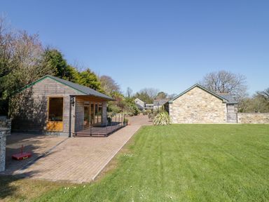 A backyard with a wooden shed and stone building next to a paved pathway and grassy lawn at Kingcup Cottage in St Columb Major
