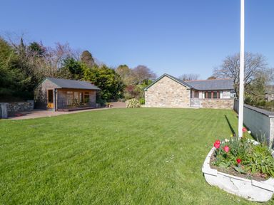 A garden with green grass and red flowers near a stone building and a wooden shed at Kingcup Cottage in St Columb Major