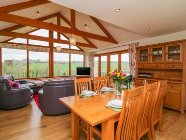 A dining area with a wooden table set for six and a living area with leather sofas and large windows showing a countryside view at Kingcup Cottage in St Columb Major