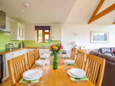 A kitchen with green tiles and wooden cabinets next to a wooden dining table set with plates glasses and a vase of flowers at Kingcup Cottage in St Columb Major