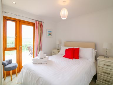 A bedroom with a bed white linens red pillows bedside tables lamps a chair and wooden doors with striped curtains at Kingcup Cottage in St Columb Major