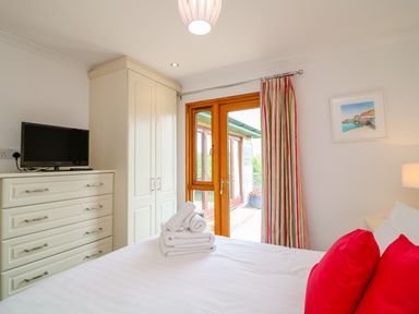 A bedroom with white bed and red pillows next to wardrobe and chest of drawers with television at Kingcup Cottage in St Columb Major