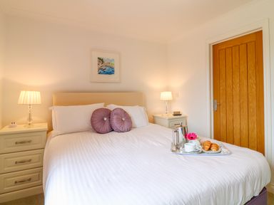 A bedroom with a bed with white sheets and purple pillows two bedside tables with lamps and a wooden door at Kingcup Cottage in St Columb Major
