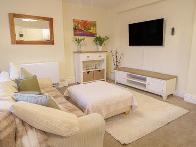 A living room with a sofa and ottoman a TV mounted on the wall and a cabinet with plants and decorations at Beside The Sea - In Town in Llandudno