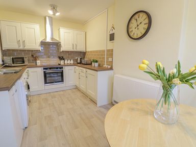 A kitchen with white cabinets brown tiled backsplash and a wooden table with yellow flowers in a vase at Beside The Sea - In Town in Llandudno