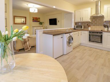 A kitchen with a round wooden table holding a vase of yellow and white tulips and an open view to a living room with a sofa and wall-mounted television at Beside The Sea - In Town in Llandudno