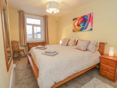 A bedroom with a double bed bedside tables a wooden chair a window with curtains and a colorful painting at Beside The Sea - In Town in Llandudno
