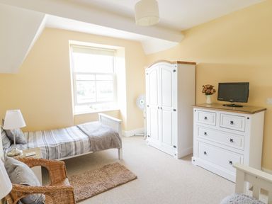 A bedroom with a single bed a wicker chair a white wardrobe a white chest of drawers a television and a window at Angorfa in Llanbedr