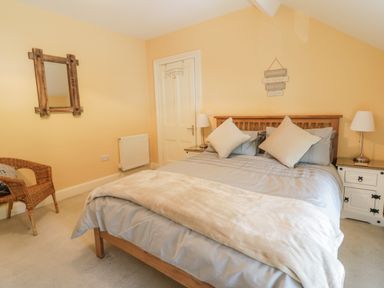A bedroom with a double bed with pillows and blanket two bedside tables with lamps a wicker chair and a wall mirror at Angorfa in Llanbedr