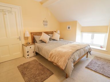 A bedroom with a wooden bed with grey bedding beige blankets and pillows two white nightstands with lamps a beige carpet and a window with a white frame at Angorfa in Llanbedr