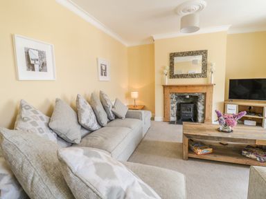 A living room with a grey sofa and multiple cushions a wooden coffee table with flowers a stone fireplace and a television on a stand at Angorfa in Llanbedr