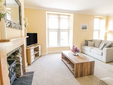 A living room with a beige sofa wooden coffee table flowers on the table a fireplace and a television at Angorfa in Llanbedr