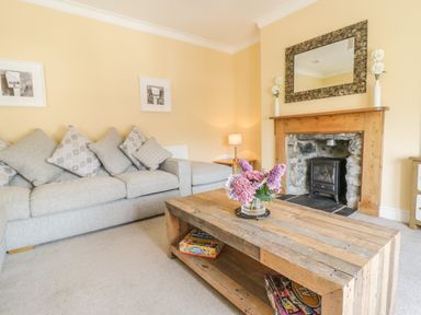 A living room with a grey sofa and patterned cushions a wooden coffee table with flowers and board games a stone fireplace with a wooden mantle and mirror at Angorfa in Llanbedr