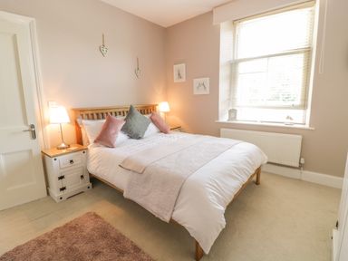 A bedroom with a wooden bed frame a white bedside table with a lamp cushions on the bed and a window with blinds at Angorfa in Llanbedr