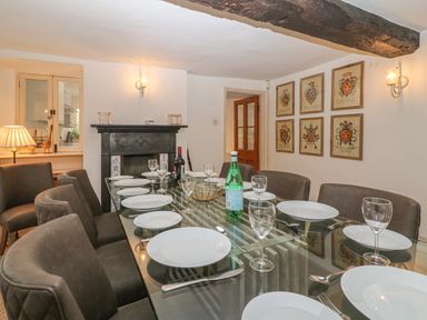 A dining room with a glass table set with white plates and glassware surrounded by chairs and a fireplace on the wall with framed pictures at Dolphin House in Burford
