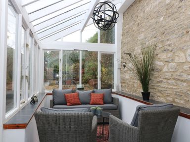A glass conservatory with wicker furniture and cushions near a stone wall at Dolphin House in Burford