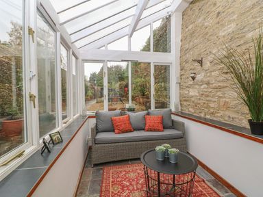 A sunroom with a grey wicker sofa with cushions a round black metal table with plants and a red patterned rug at Dolphin House in Burford