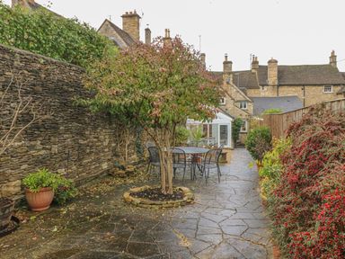 A stone paved garden with a tree and outdoor table and chairs beside a stone wall and bushes at Dolphin House in Burford