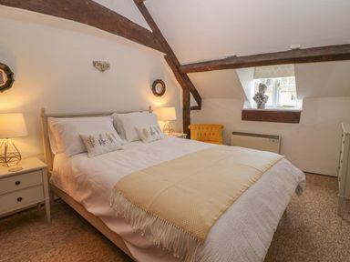 A bedroom with a bed white bedding two bedside tables with lamps an orange chair under a window and exposed wooden beams at Dolphin House in Burford