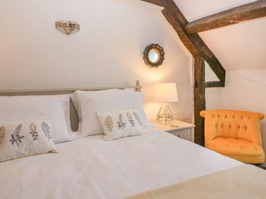 A bedroom with a bed with white bedding pillows with plant designs a bedside table with a lamp a small mirror on the wall and an orange tufted chair with wooden beams in Burford