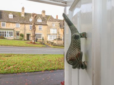 A fish shaped door knocker on a white door with stone buildings and grass across the road at Dolphin House in Burford
