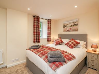 A bedroom with a bed featuring red plaid pillows and blanket, bedside tables with lamps, a window with red plaid curtains and a landscape painting above the bed at Tilly's Cottage in Edington