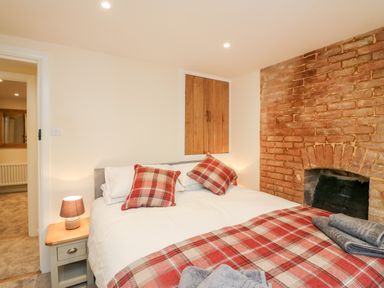 A bedroom with a double bed with plaid pillows and blanket a bedside table with lamp and an exposed brick wall at Tilly's Cottage in Edington