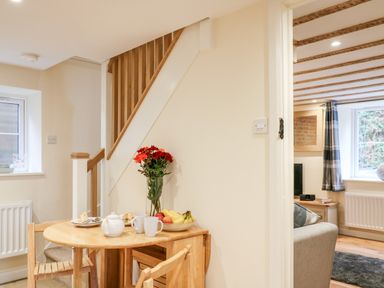 A dining area with a small wooden table set with tea and fruit beside stairs and a view into a living room at Tilly's  Cottage in Edington