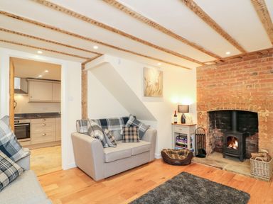 A living room with a sofa, pillows, a wood burning stove in a brick fireplace, a small table with a lamp and books and a view into a kitchen at Tilly's Cottage in Edington