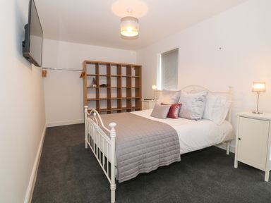 A bedroom with a white metal bed a gray quilt and pillows bedside tables with lamps a wooden shelving unit and a wall-mounted television at 8 Marina View in Pwllheli