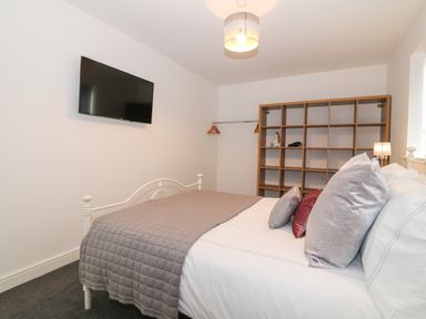 A bedroom with a bed covered by a gray quilt and several pillows a wooden shelf unit with an iron and clothes hangers on a rail and a wall-mounted television at 8 Marina View in Pwllheli