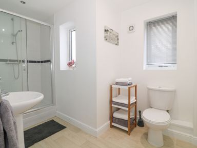 A bathroom with a shower enclosure a sink a toilet a wooden shelf with folded towels and two windows at 8 Marina View in Pwllheli