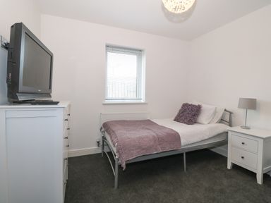 A bedroom with a bed with purple blanket and pillow a window with blinds a white chest of drawers with a tv and a white bedside table with a lamp at 8 Marina View Pwllheli