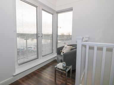A corner with large windows with blinds a grey armchair with a pillow and a small glass side table next to a white staircase railing at 8 Marina View in Pwllheli